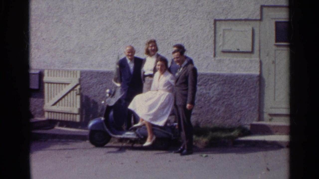 The home-movie frame shows the adults shifting slightly around the blue scooter, with the exterior wall and doorway still visible behind them.