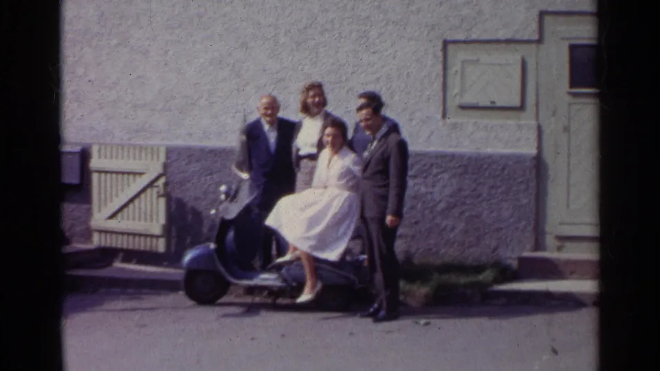 The home-movie frame shows the adults shifting slightly around the blue scooter, with the exterior wall and doorway still visible behind them.