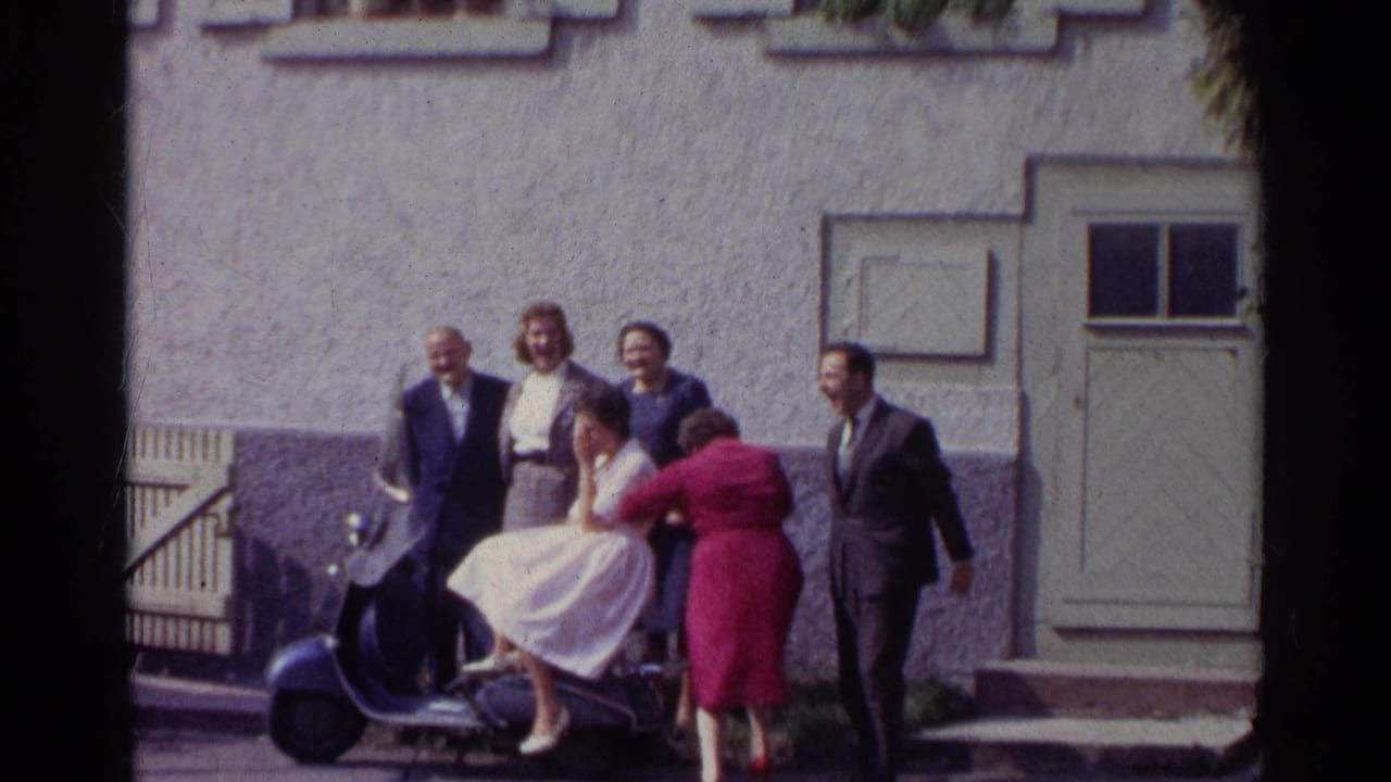 A fifth adult wearing a red coat enters or adjusts the group pose beside the seated woman on the scooter in the Germany 1961 street scene.