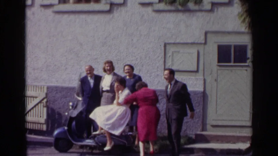 A fifth adult wearing a red coat enters or adjusts the group pose beside the seated woman on the scooter in the Germany 1961 street scene.