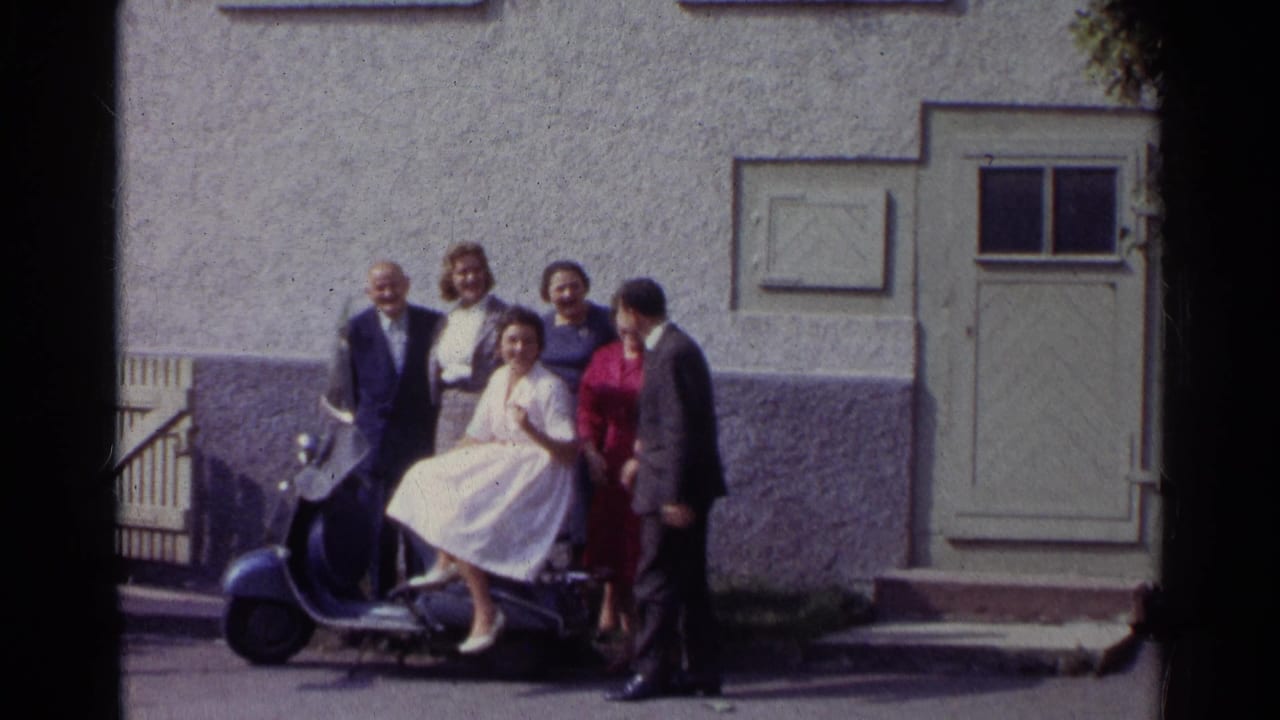 In 1961 Germany, well-dressed adults gather around a woman in a white dress on a blue scooter outside a stucco facade.