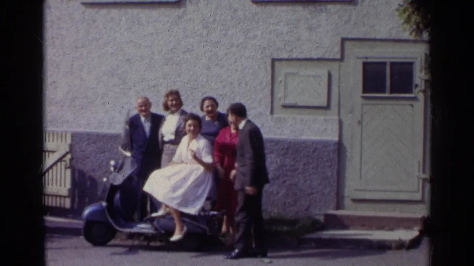 In 1961 Germany, well-dressed adults gather around a woman in a white dress on a blue scooter outside a stucco facade.