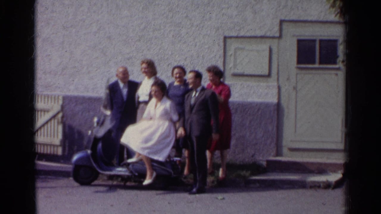 Well-dressed adults in 1961 Germany stand and wave around a woman on a blue scooter outside a stucco building.