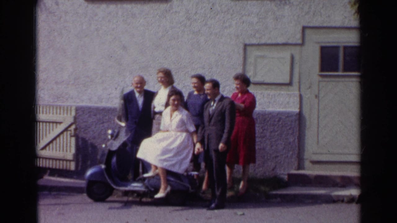 A 1961 German home-movie frame shows adults posed tightly around a blue scooter in front of a shuttered door and stucco wall.
