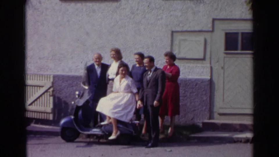 A 1961 German home-movie frame shows adults posed tightly around a blue scooter in front of a shuttered door and stucco wall.