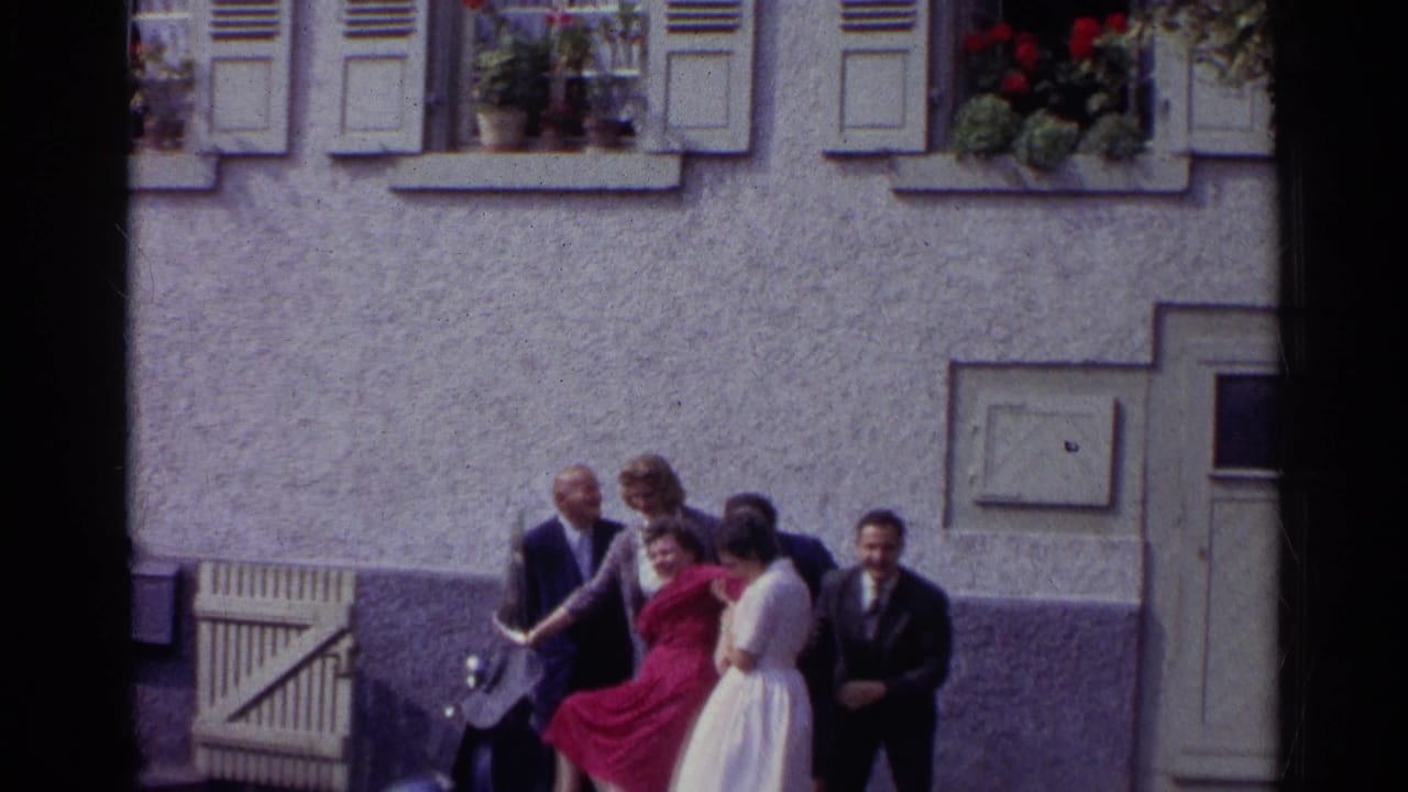 The group moves and waves near the scooter beneath shuttered windows and flower boxes on a German building facade.