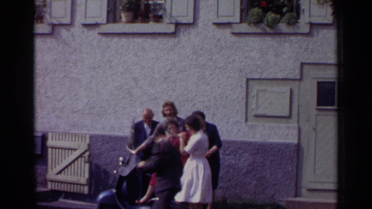 In 1961 Germany, adults in suits and dresses cluster around a dark scooter in a courtyard as one woman is lifted or posed near the seat.