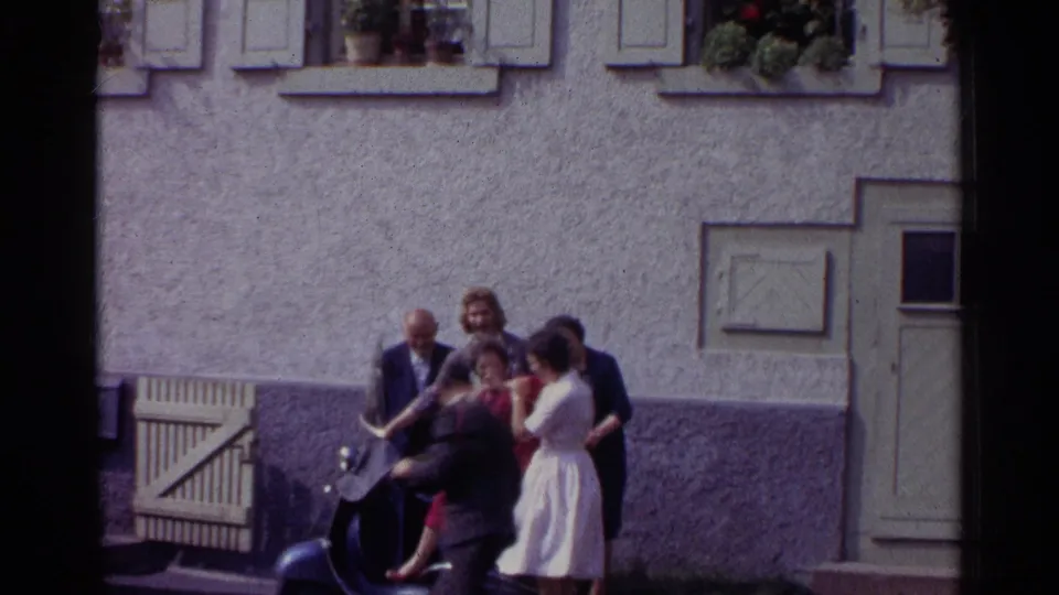In 1961 Germany, adults in suits and dresses cluster around a dark scooter in a courtyard as one woman is lifted or posed near the seat.
