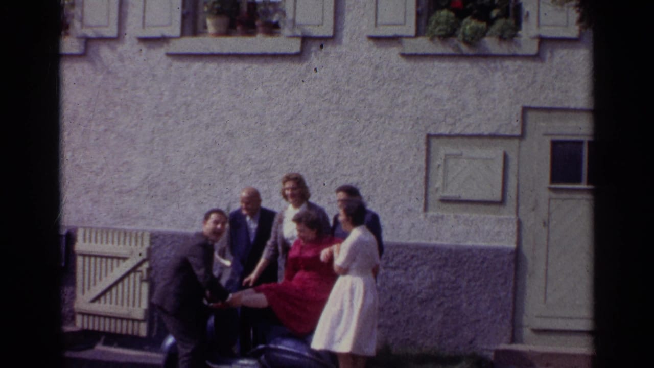 In 1961 Germany, a woman in a red dress is held or steadied on a dark scooter while several adults smile and pose beside a stucco building.
