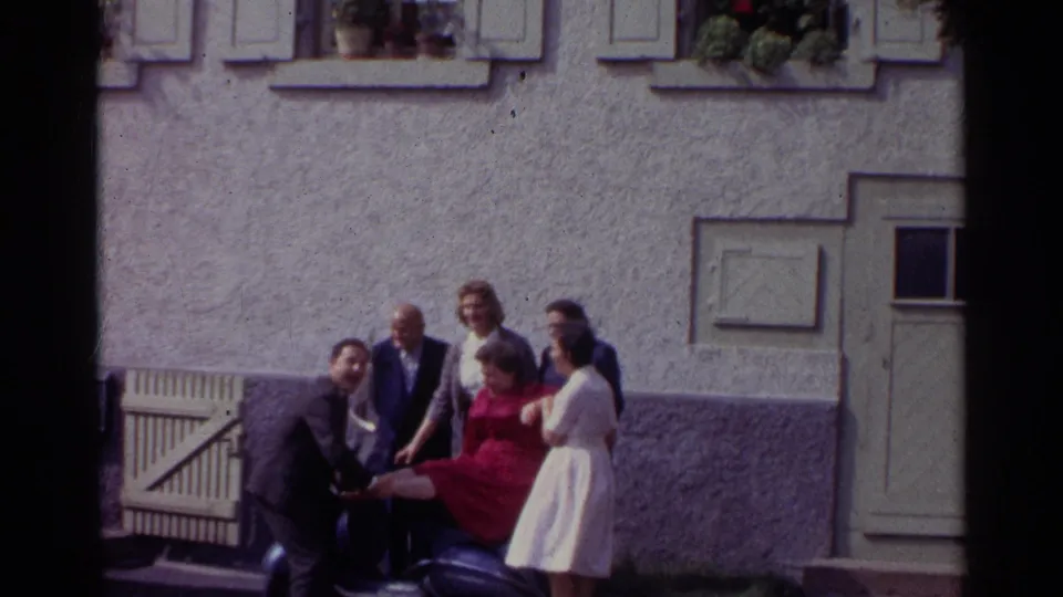 In 1961 Germany, a woman in a red dress is held or steadied on a dark scooter while several adults smile and pose beside a stucco building.
