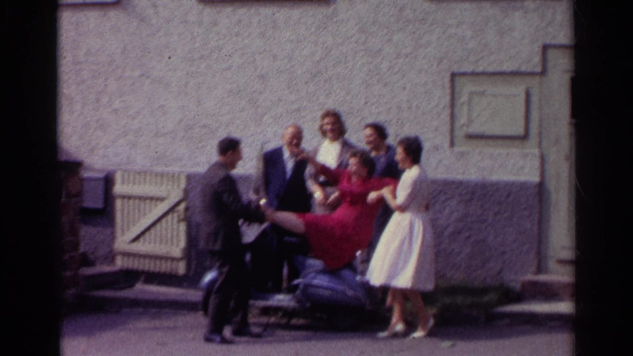 In 1961 Germany, a man in a dark suit helps hold a woman in a red dress on the scooter as other adults stand nearby in the courtyard.