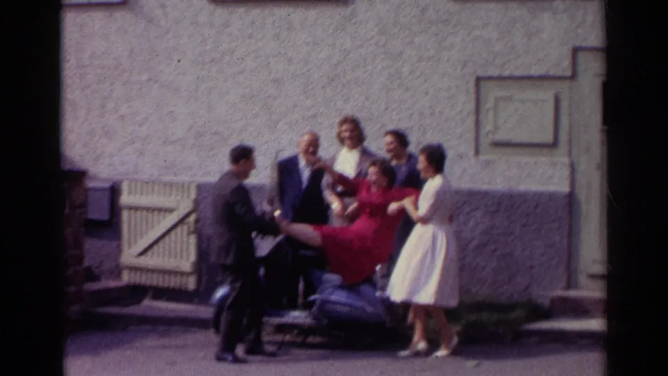 In 1961 Germany, a man in a dark suit helps hold a woman in a red dress on the scooter as other adults stand nearby in the courtyard.