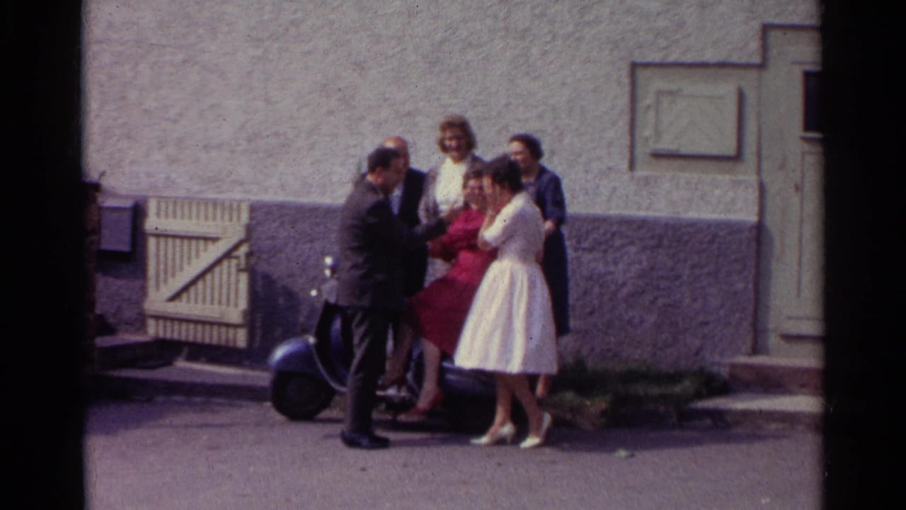 In Germany 1961 home-movie footage, adults stand outside a stucco building near a scooter, including women in red and pale dresses.