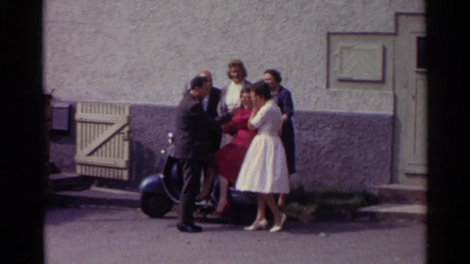 In Germany 1961 home-movie footage, adults stand outside a stucco building near a scooter, including women in red and pale dresses.