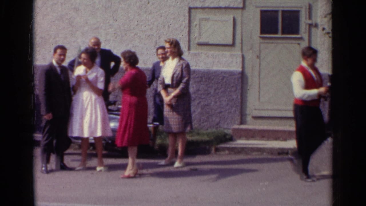Germany 1961 frame showing adults outside a building by a parked car, with formal clothing and a red-waistcoated man at frame right.