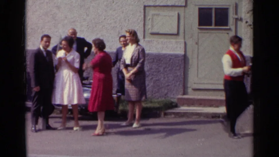 Germany 1961 frame showing adults outside a building by a parked car, with formal clothing and a red-waistcoated man at frame right.