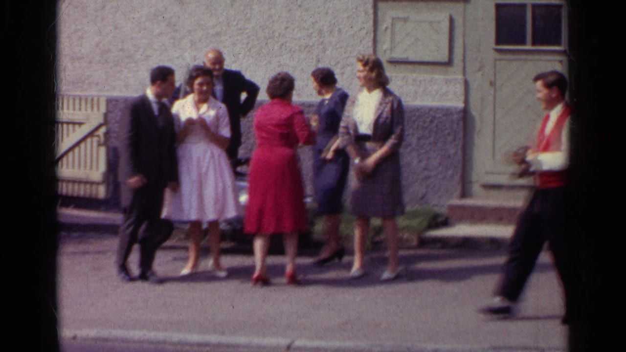 A German 1961 home-movie frame shows adults conversing in the street while a red-waistcoated man crosses the foreground.