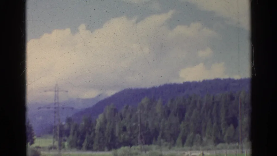 A brief Germany 1961 landscape view shows a wooded hillside beneath large clouds, with fields and a distant utility pylon.