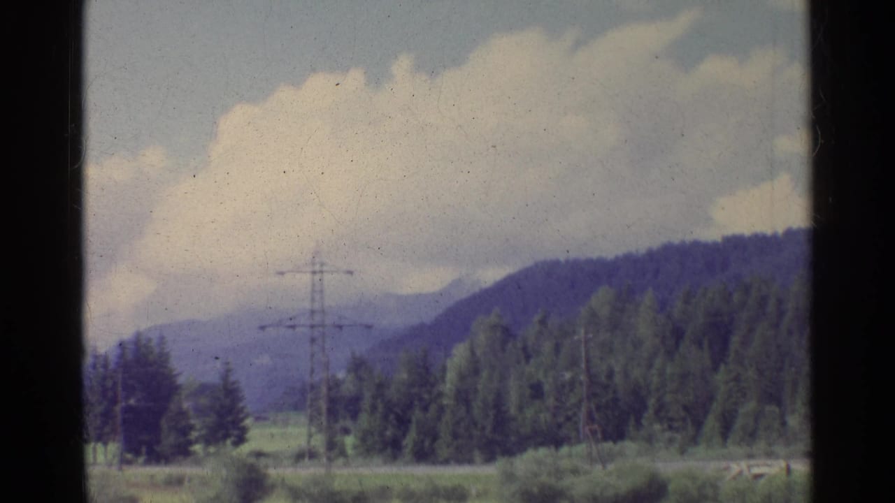 Forested hillside, open fields, distant mountains, clouds, and power pylons in a rural view.