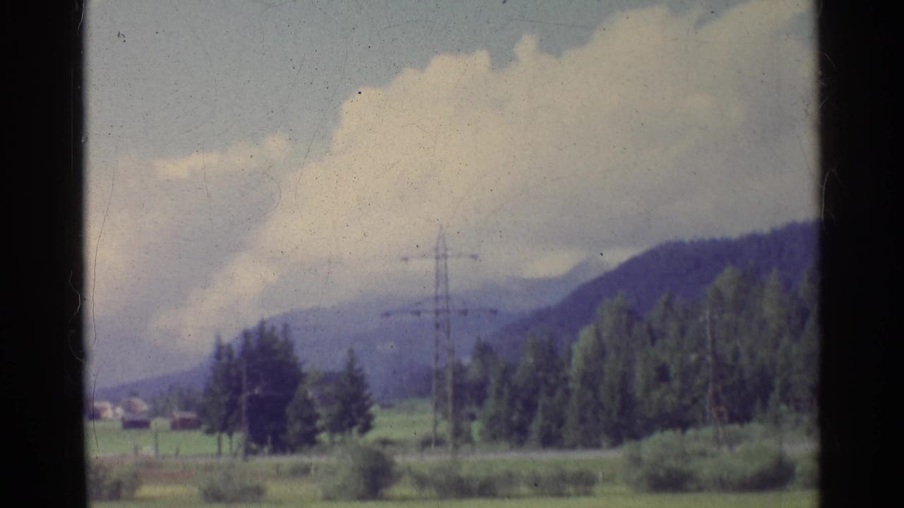 1961 German rural view with low vegetation, a pylon, forested slopes, clouds, and small distant houses near the horizon.