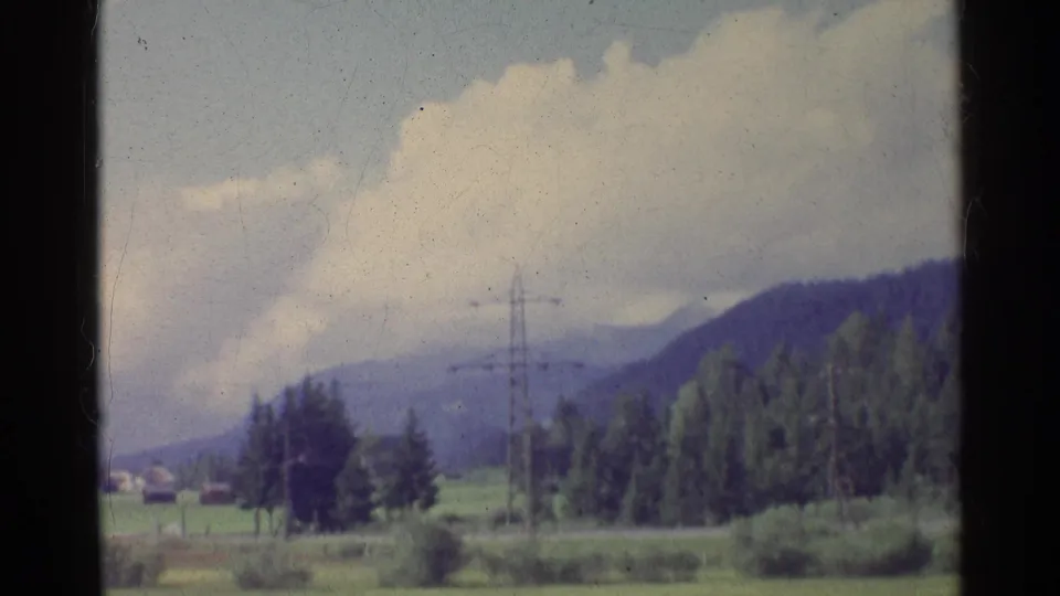 1961 German rural view with low vegetation, a pylon, forested slopes, clouds, and small distant houses near the horizon.