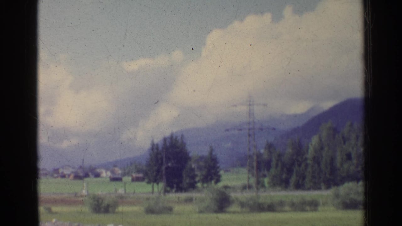 1961 German countryside with open fields, distant houses, trees, electricity pylons, wooded hills, and large white clouds.
