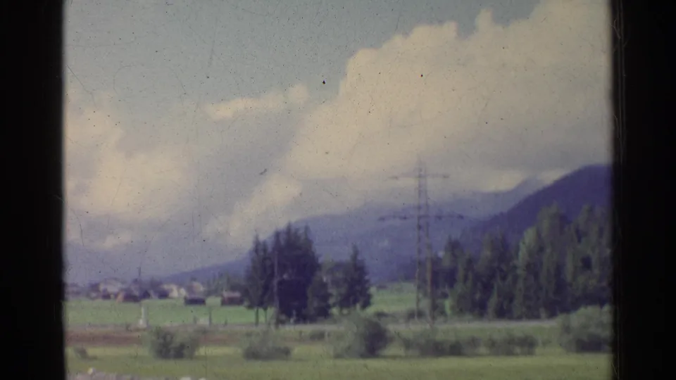 1961 German countryside with open fields, distant houses, trees, electricity pylons, wooded hills, and large white clouds.