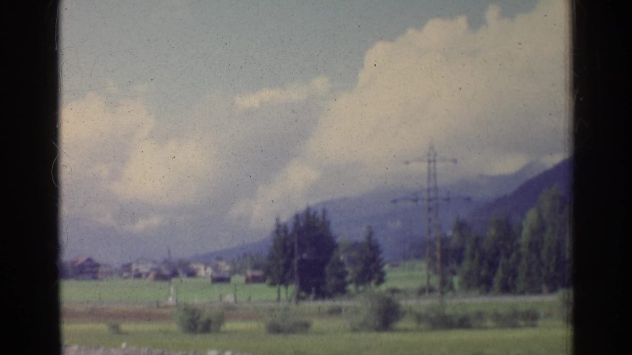 1961 German countryside view with fields, distant houses, electricity pylons, tree clusters, and mountains under a cloudy sky.