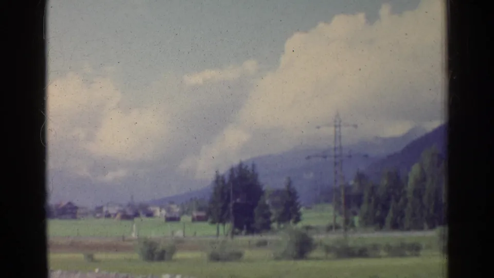 1961 German countryside view with fields, distant houses, electricity pylons, tree clusters, and mountains under a cloudy sky.