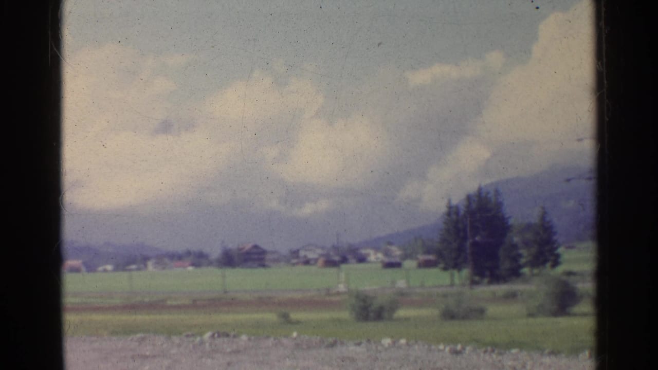 A moving 8mm view of the German countryside shows a blurred foreground, green farmland, distant houses, trees, and mountains.