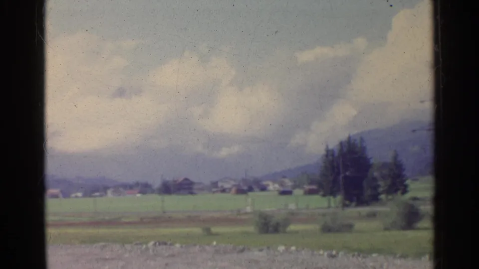 A moving 8mm view of the German countryside shows a blurred foreground, green farmland, distant houses, trees, and mountains.