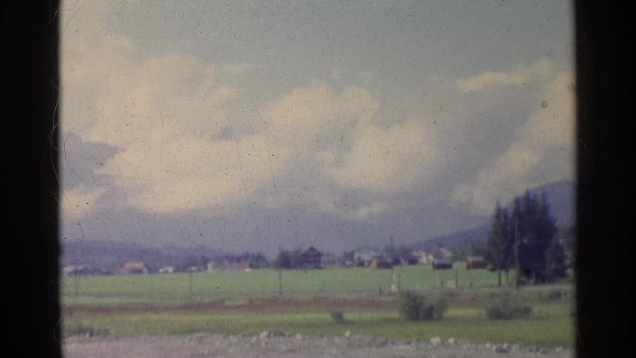 The 1961 home-movie frame looks across German fields toward distant village buildings, a tree line, pylons, and mountains.