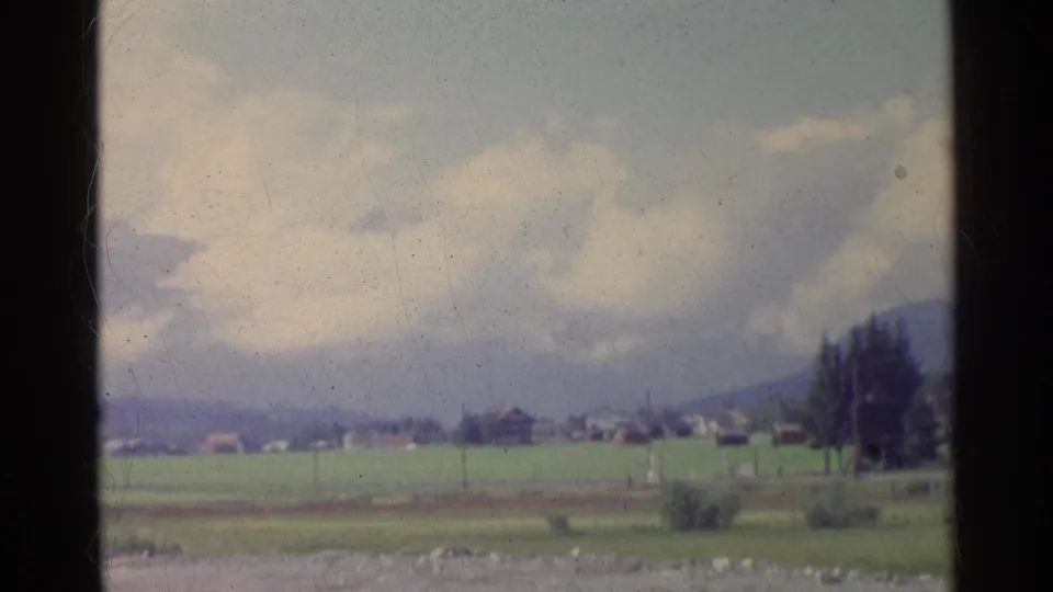 The 1961 home-movie frame looks across German fields toward distant village buildings, a tree line, pylons, and mountains.