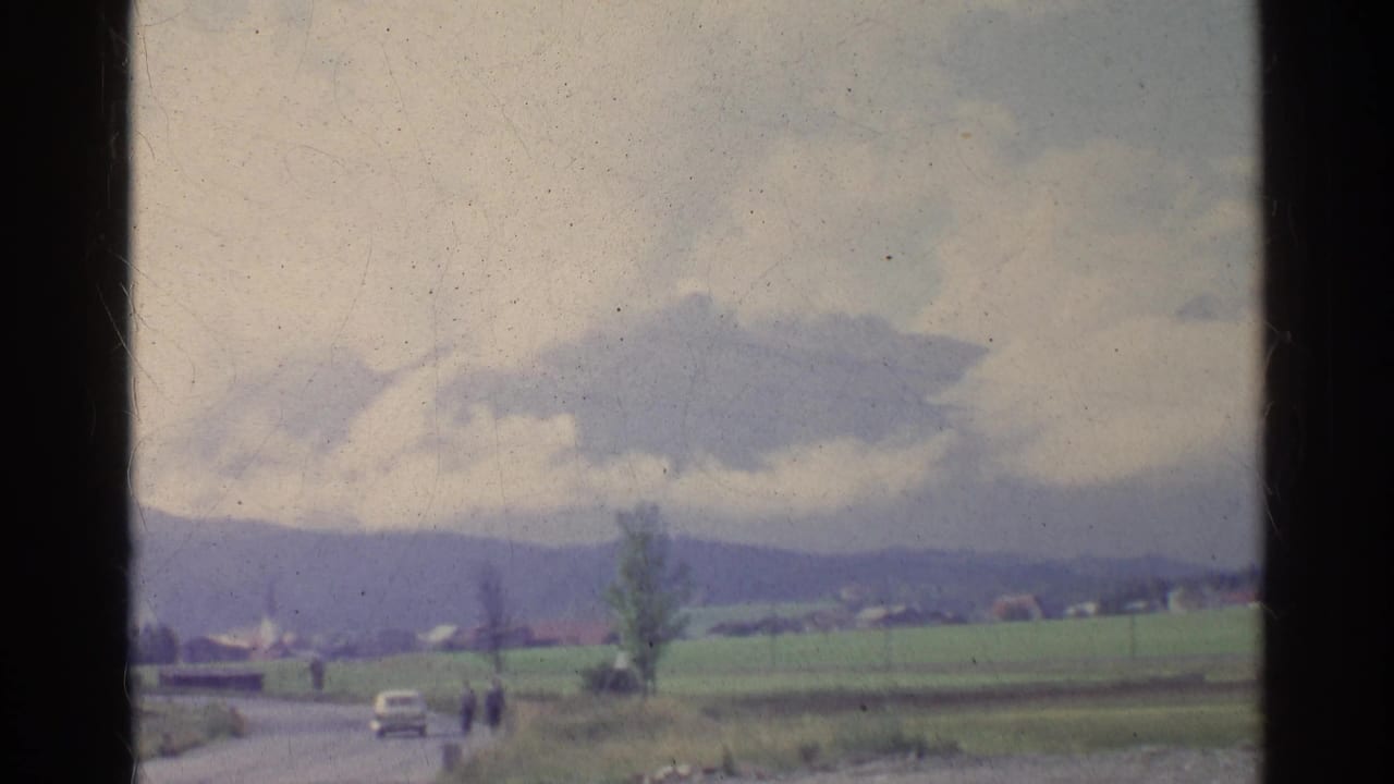 1961 German countryside road scene with a small car, distant walking figures, fields, hills, and a cloudy sky viewed from a moving vehicle.