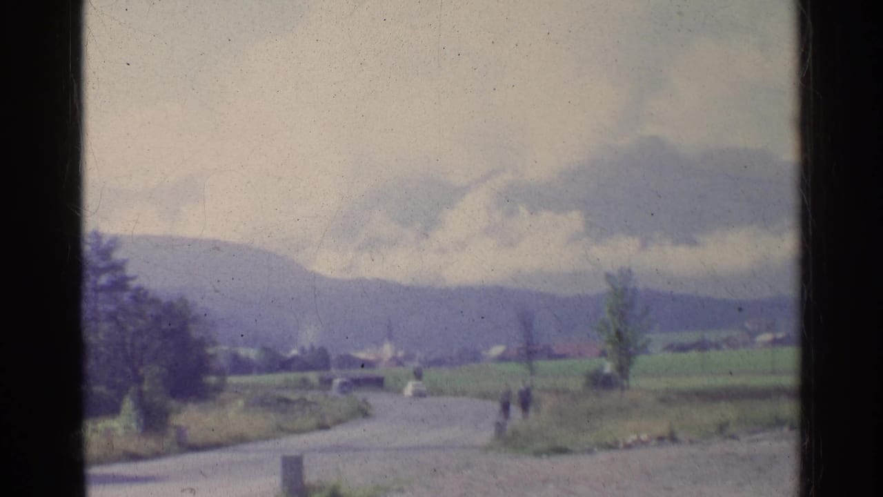 1961 German landscape with a curving rural road, small distant pedestrians, green fields, low hills, and a cloudy sky.