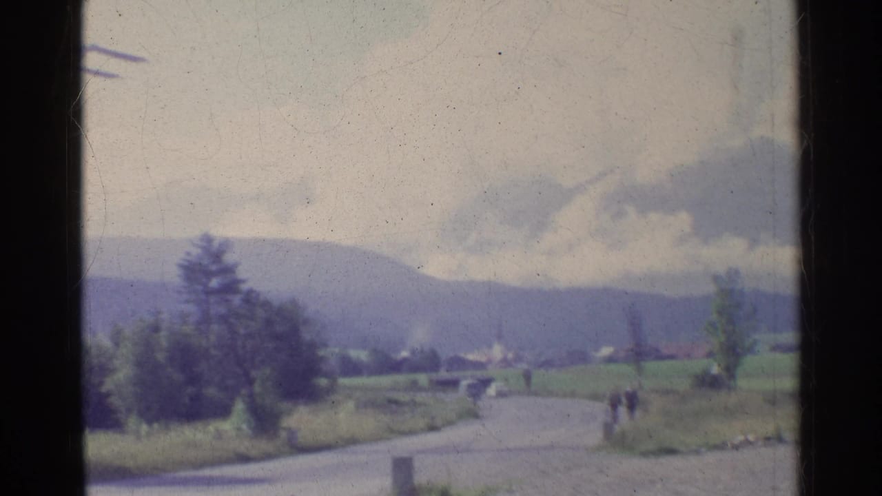 1961 Germany 8mm view of a countryside road, distant hills, trees, small structures, and two far-off figures framed by dark film borders.