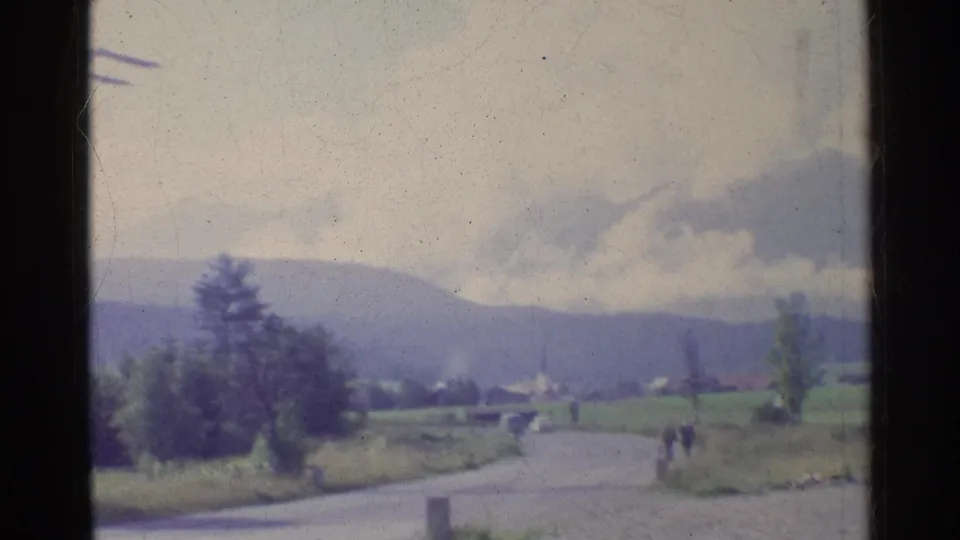 1961 Germany 8mm view of a countryside road, distant hills, trees, small structures, and two far-off figures framed by dark film borders.