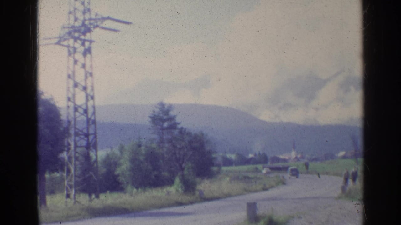 Roadside landscape with an electricity pylon, distant hills, trees, a car, and small figures near the road.