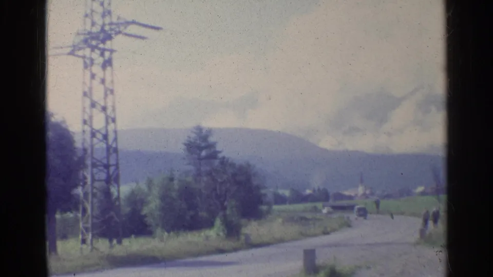 1961 Germany rural roadside scene with a prominent electricity pylon, mountain backdrop, trees, a car, and distant pedestrians.
