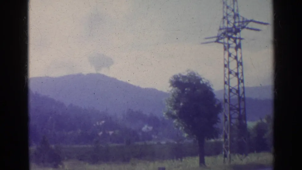 Faded 1961 Germany countryside frame showing wooded mountains, open fields, a lone tree, and an electricity pylon near the foreground.