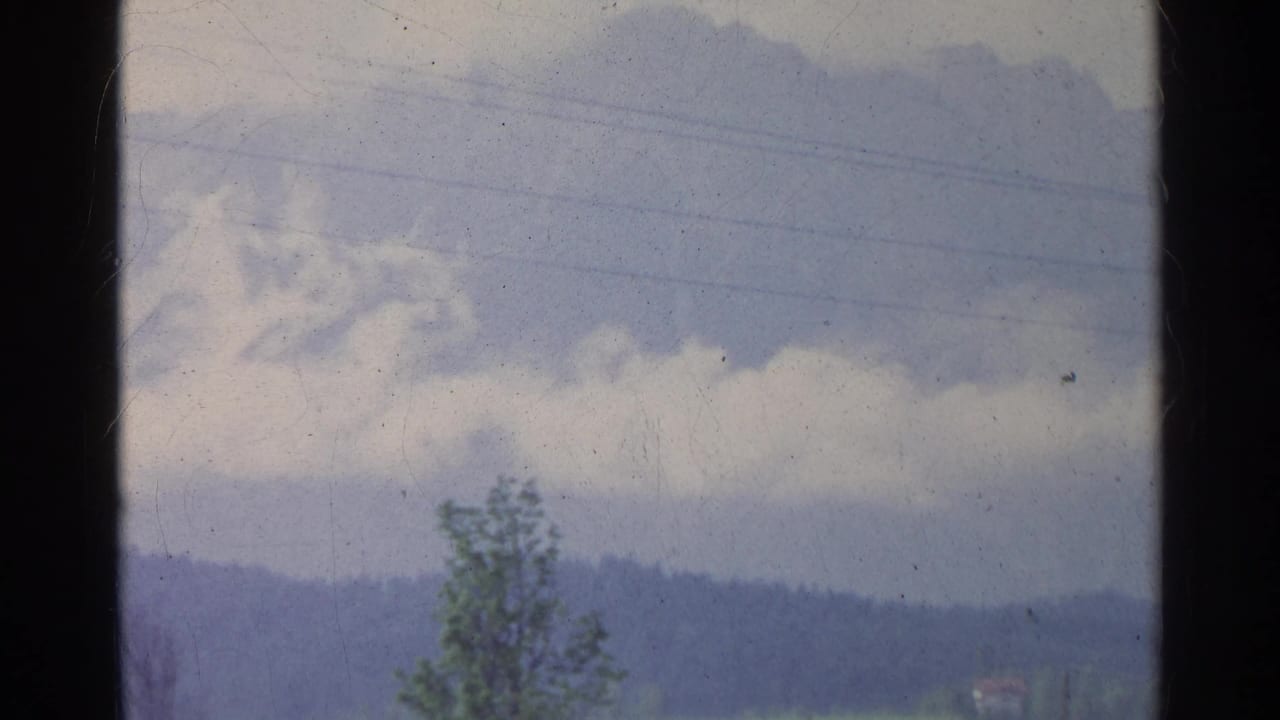 German 8mm home-movie frame showing a large cloud formation above distant hills, with a foreground tree and faint lines in the sky.
