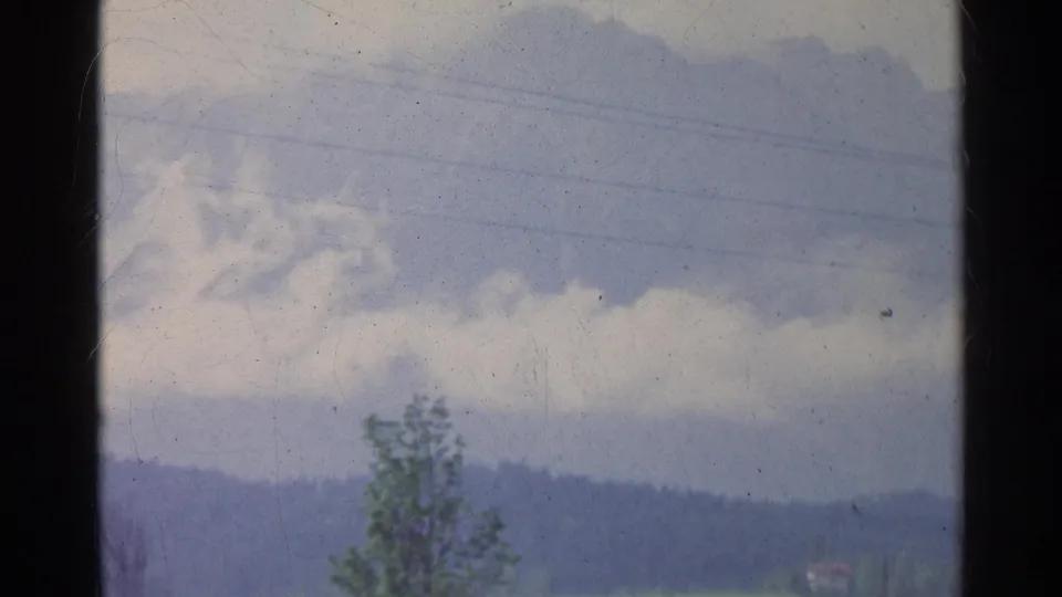 German 8mm home-movie frame showing a large cloud formation above distant hills, with a foreground tree and faint lines in the sky.