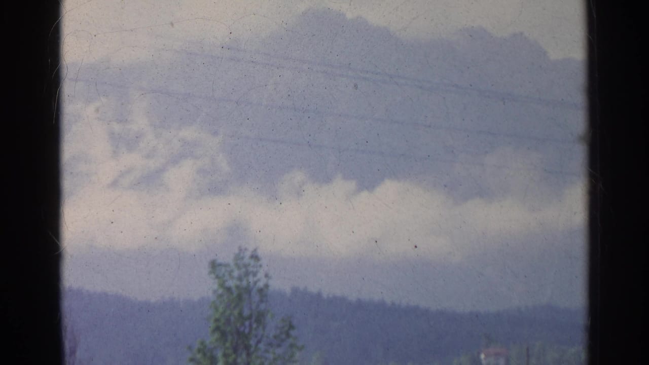 1961 Germany home-movie frame with horizontal power lines, overcast sky, a lone tree, and hazy hills in the background.