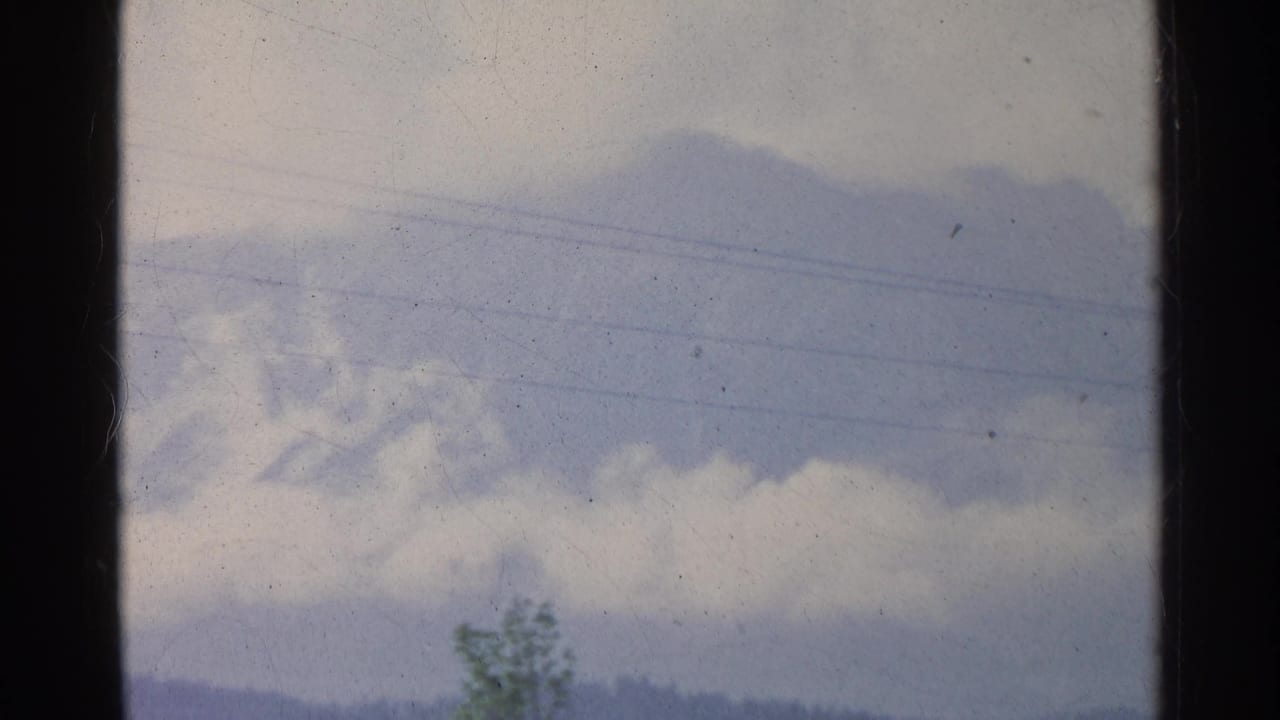 1961 Germany 8mm frame showing a cloudy sky crossed by faint power lines, with a tree and distant wooded hills below.