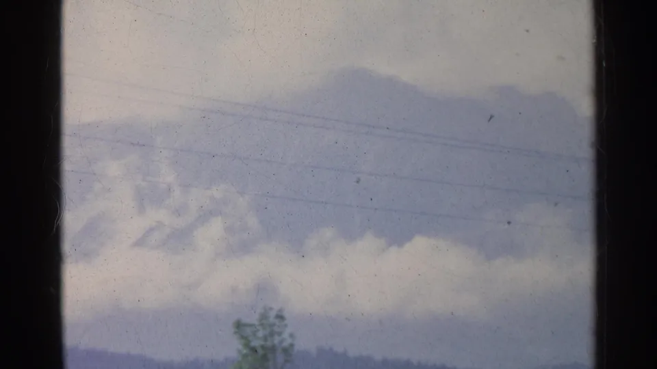 1961 Germany 8mm frame showing a cloudy sky crossed by faint power lines, with a tree and distant wooded hills below.