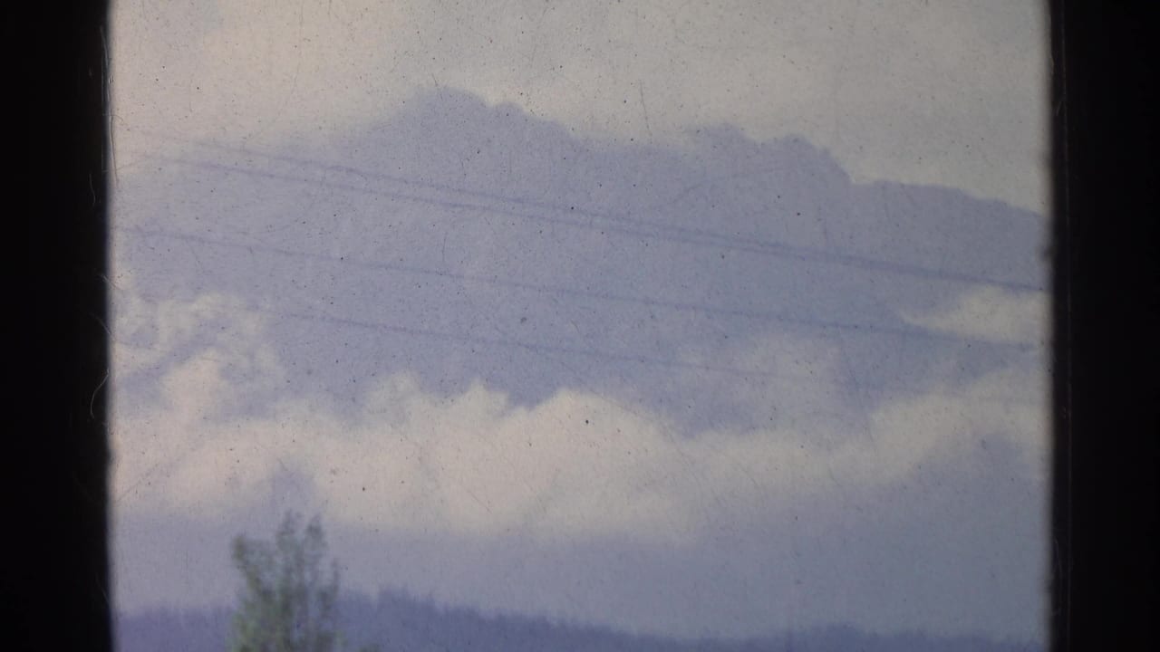 1961 German home-movie frame showing hazy mountains, cloud, and a small band of trees in soft 8mm color.