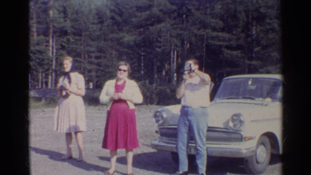 Three adults stand beside a white sedan on a sunny roadside near pine trees.
