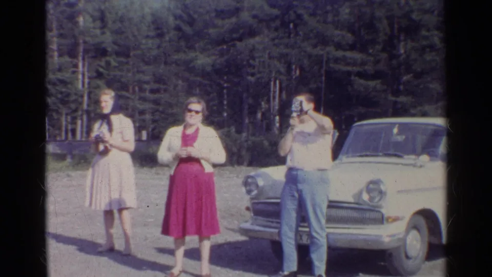 Three adults appear beside a white mid-century sedan on a German roadside, with two people holding cameras near a pine forest.