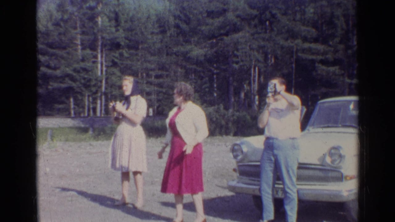 1961 German roadside frame with three adults, handheld cameras, a white sedan, and dense trees behind them.