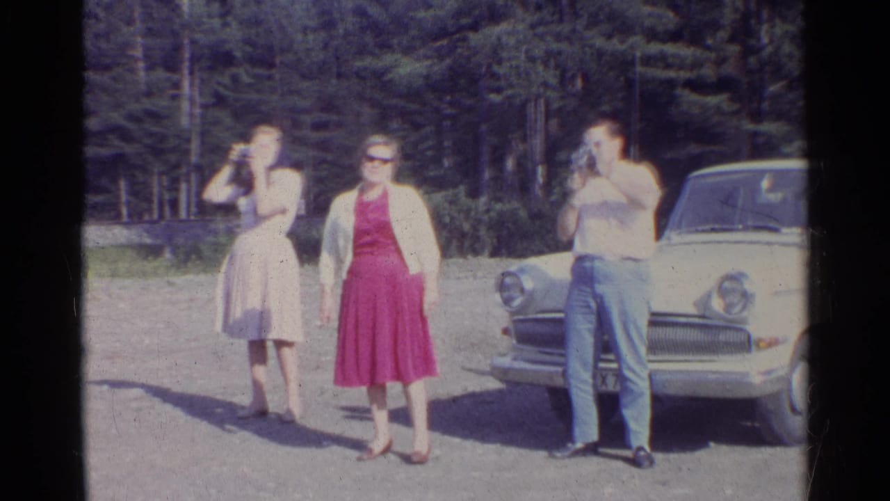 A woman in a pink dress and two companions stand beside a white sedan during a 1961 German road trip stop.